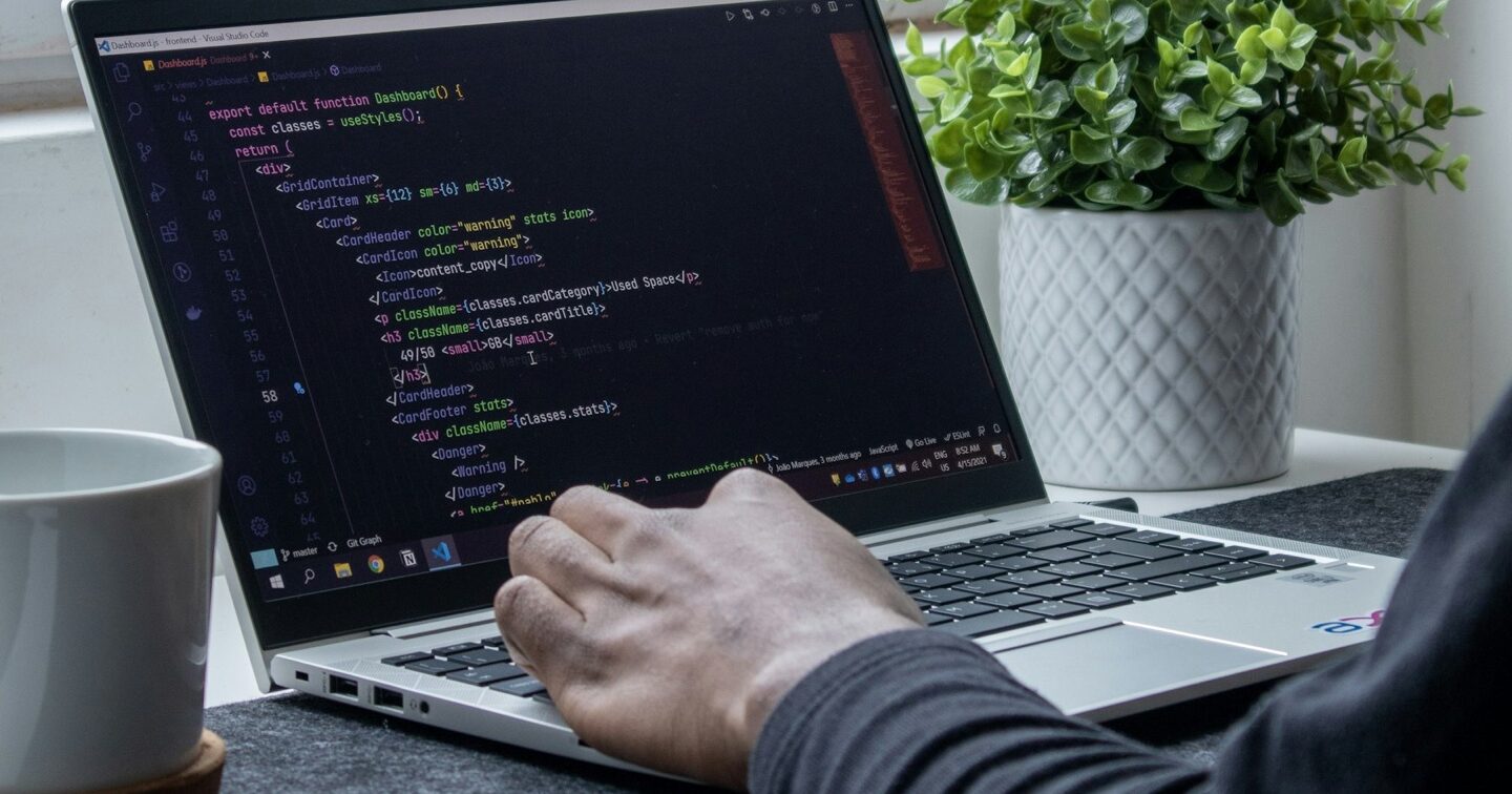 Developer typing code on a laptop at a window desk with a coffee mug and potted plant