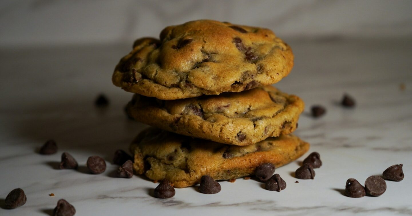A stack of three chocolate chip cookies on a white marble surface scattered with chocolate chips