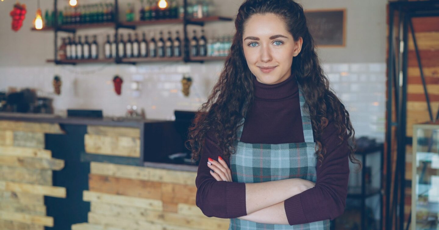 Smiling owner in an apron standing with arms folded in front of the counter of her small coffee shop