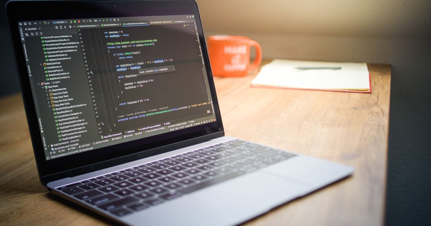 Laptop on a wooden desk showing code in an editor, with a mug and notebook behind
