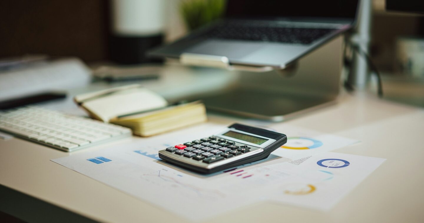 Calculator and printed budget charts on an office desk beside a laptop