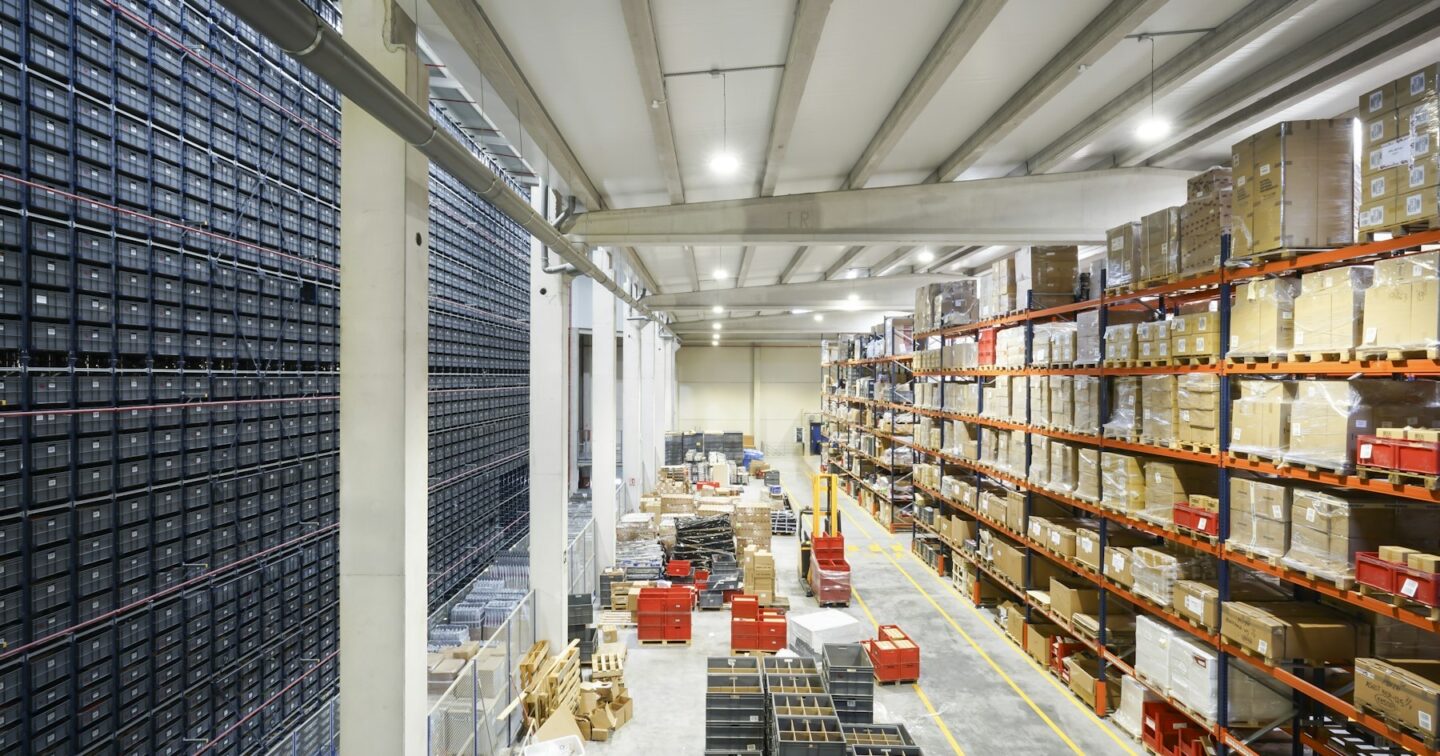 Long warehouse aisle with tall orange racking stacked with cardboard boxes and pallets beside an automated storage wall