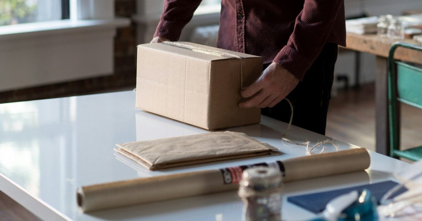 Person in a burgundy shirt packing a cardboard parcel at a white table with kraft paper and twine in a bright small-business workspace