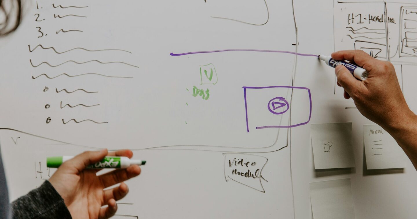 Two people sketching web app wireframes and page layouts on a whiteboard with marker pens