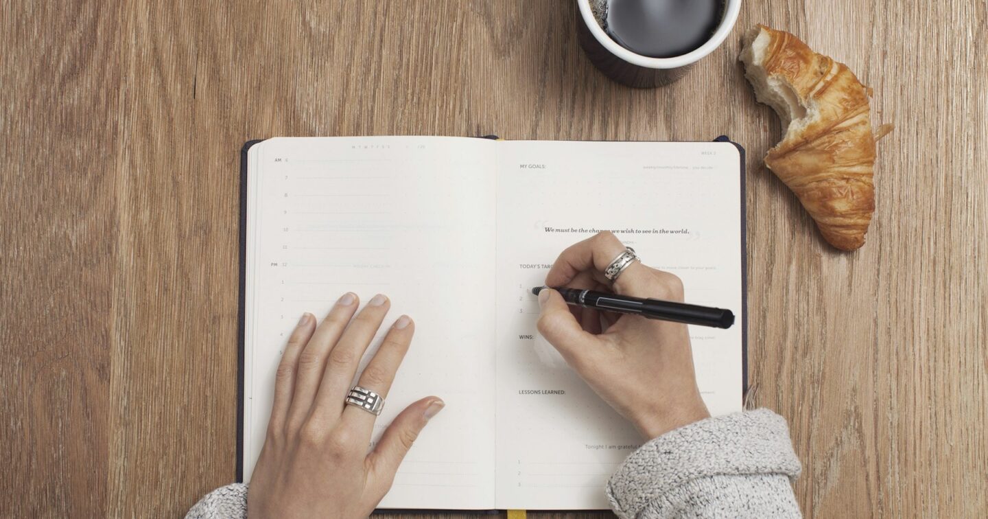 Overhead view of hands writing goals in an open planner on a wooden desk beside a cup of coffee