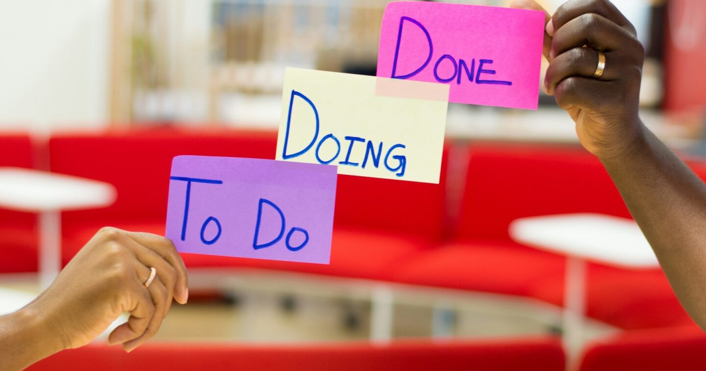 Two hands holding up handwritten sticky notes reading To Do, Doing and Done in a bright office