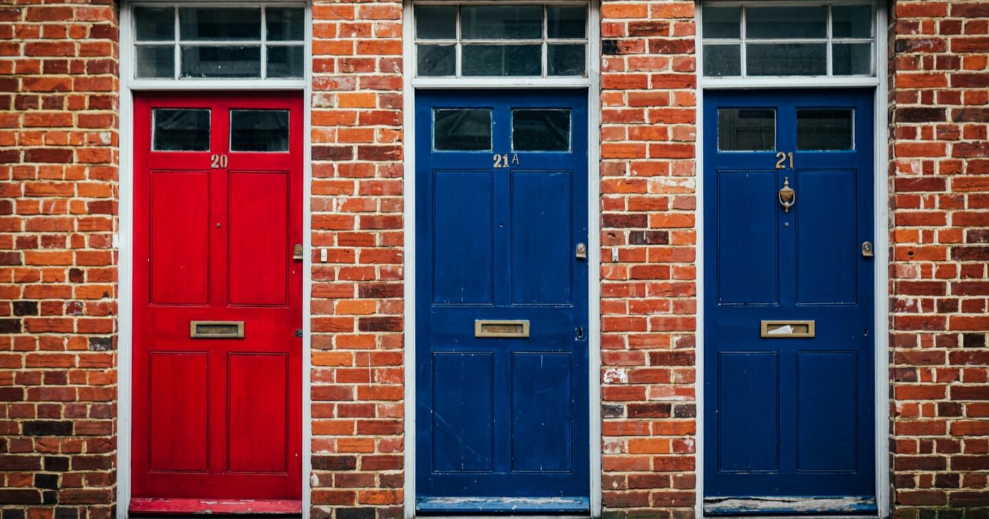 Three numbered front doors side by side on a British brick terrace, like choosing between .co.uk, .uk and .com addresses for a business domain name