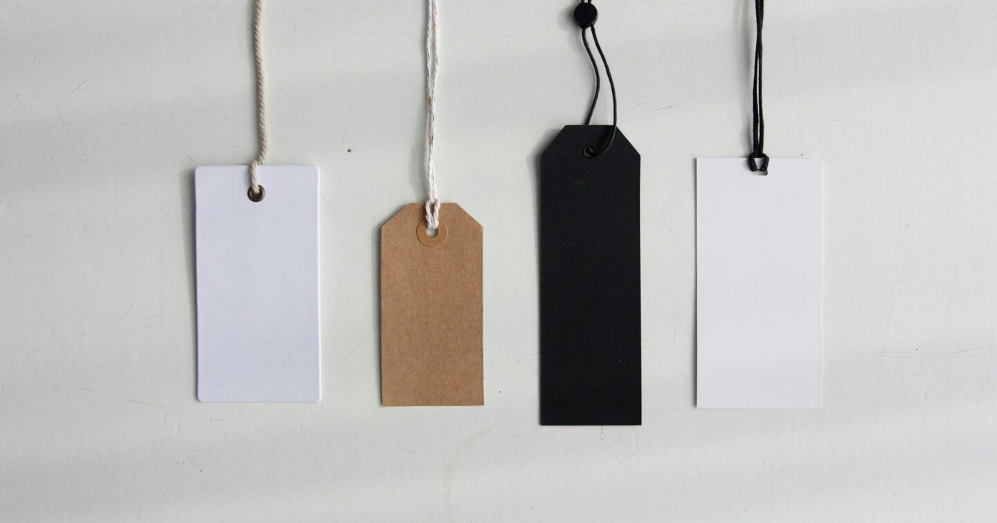 Four blank swing tags in white, brown kraft and black hanging on strings against a pale background, standing for the different price points of an ecommerce website