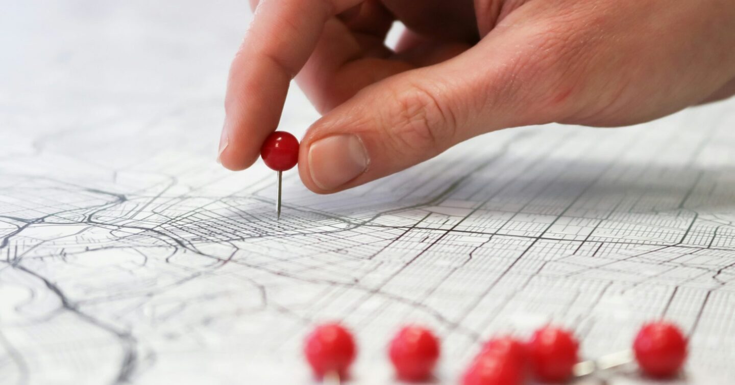 A hand pressing a red push pin into a street map, with more red pins in the foreground, like a business marking its location and service area on Google Maps