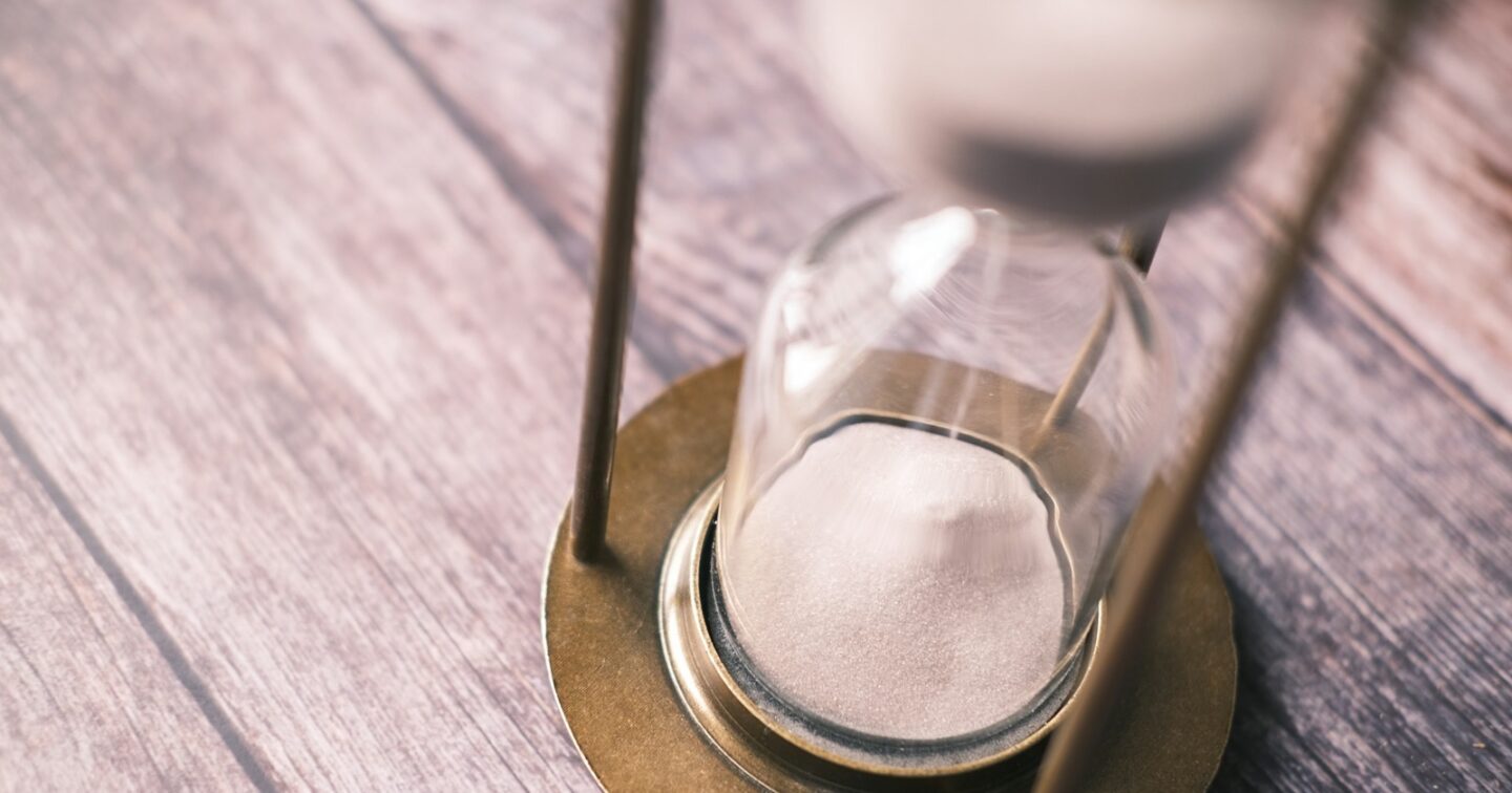 The lower half of a brass hourglass on a wooden table, with most of the sand already run through
