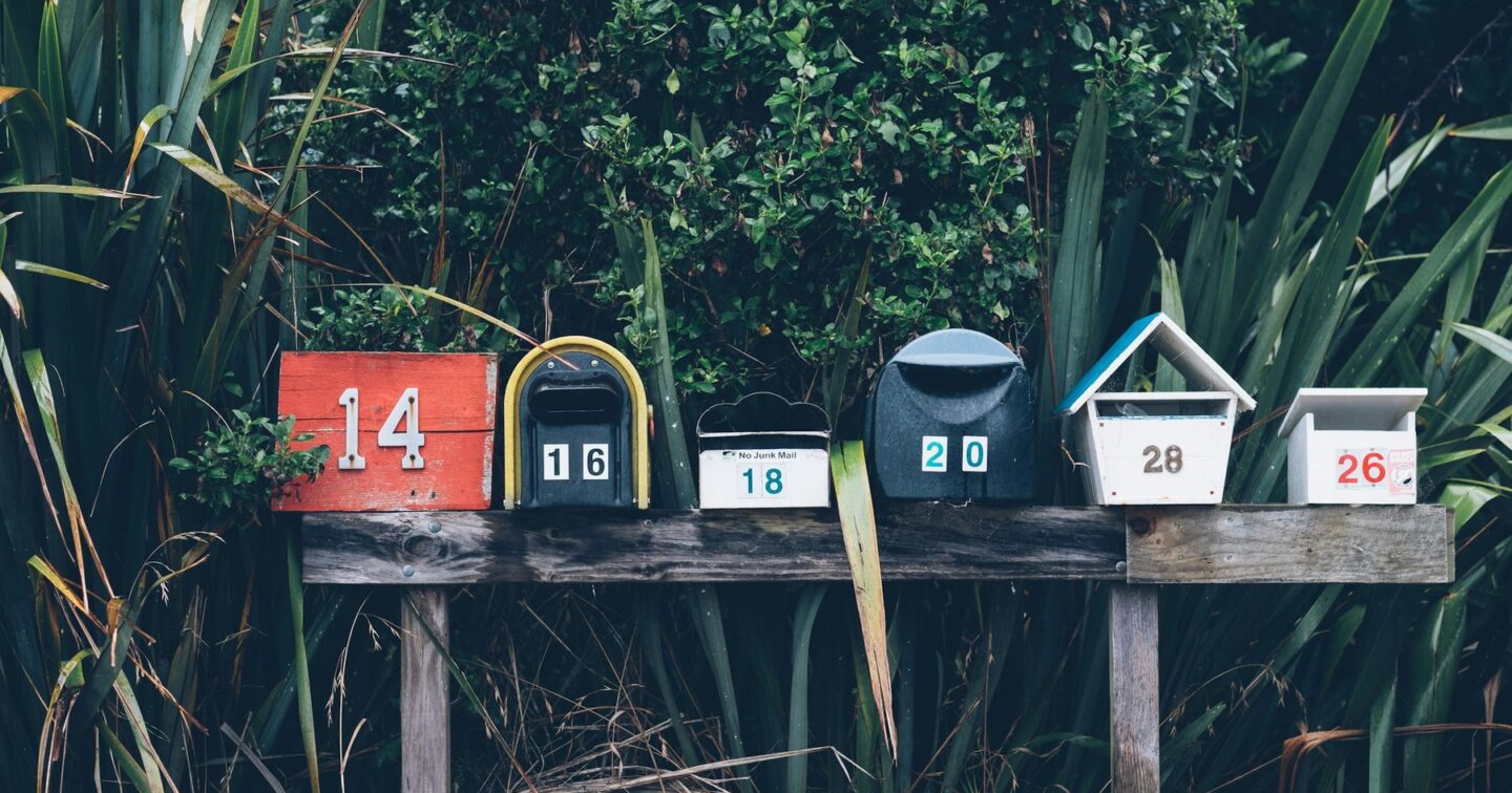A row of six numbered letterboxes in different shapes and colours on a wooden shelf in front of a hedge, one with a "No Junk Mail" label, illustrating business email reaching the right inbox