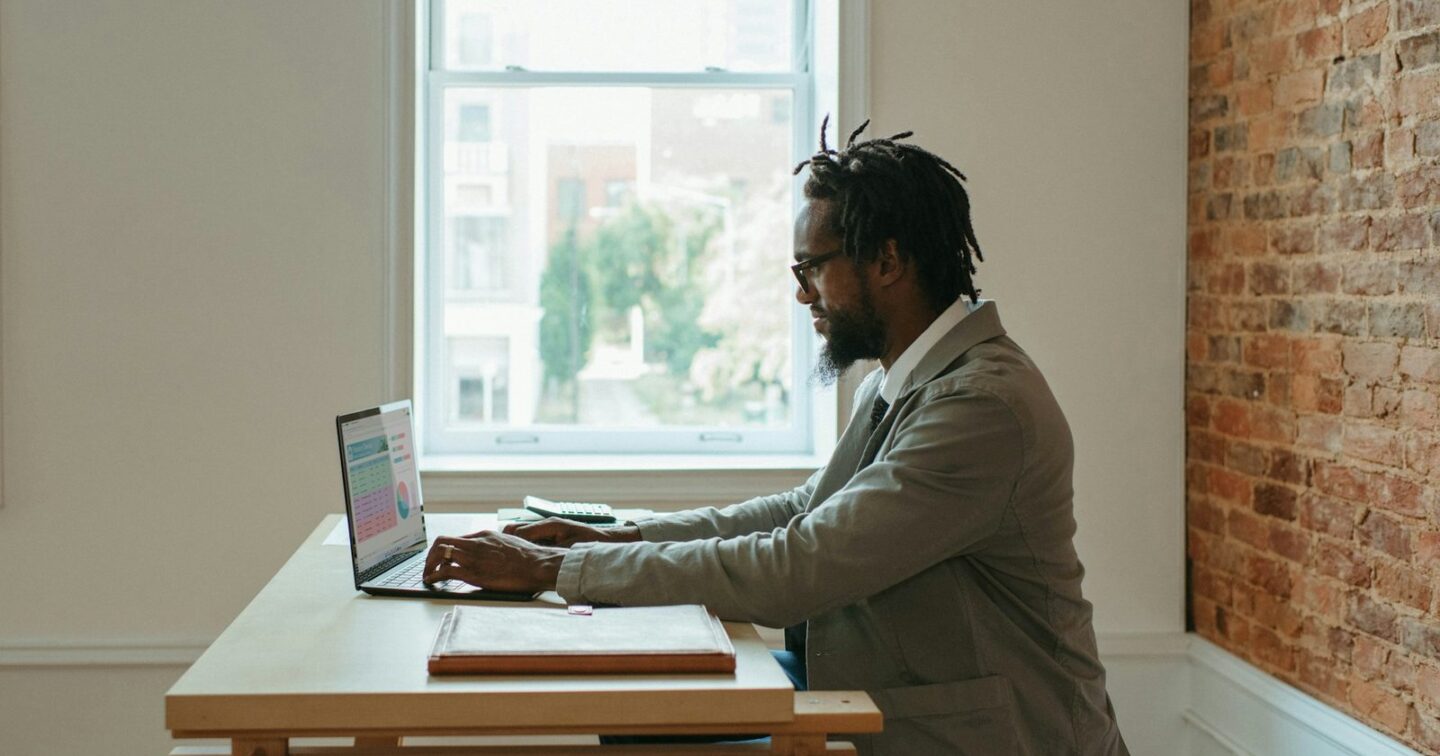 A man in a grey jacket working on a laptop at a wooden desk beside a window, with a calculator and a document folder next to him, checking the details of his business website